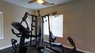 This single-floor fitness space is purpose-built for home workouts, offering a well-organized and functional environment for strength training and exercise. The room is anchored by a substantial multi-station cable machine, which features a cable crossover system with adjustable pulley attachments, allowing for a wide variety of upper and lower body exercises. A seated workout station is positioned nearby, providing dedicated support for targeted muscle training. The flooring is finished with dark rubberized gym matting, offering durability and safety during high-intensity workouts. Neutral-toned walls keep the space feeling clean and focused, while a ceiling fan overhead ensures consistent airflow and comfort during exercise sessions. A window dressed with vertical blinds allows natural light to filter into the room, brightening the space without causing glare. The overall layout is efficient and compact, making excellent use of the available footprint while accommodating a full suite of resistance training equipment. This space is well-suited for individuals seeking a comprehensive and convenient home gym experience.