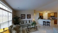 This single-story space showcases a thoughtfully designed open-concept layout that seamlessly connects the living area to the kitchen. The living room serves as the central gathering space, featuring plush, light gray upholstered seating arranged around a glass-top coffee table with ornate metal legs, anchored by a traditional area rug. Decorative touches include framed botanical artwork displayed on neutral-toned walls, potted greenery, and candlestick accents that contribute to the warm, inviting ambiance. Soft, neutral carpeting extends throughout the living area, enhancing the comfortable atmosphere.

Adjacent to the living room, the kitchen is partially visible and features warm wood cabinetry, stainless steel appliances, and a raised counter or breakfast bar accompanied by a tall bar stool. The kitchen benefits from brighter, recessed lighting that creates a welcoming contrast to the softly lit living space.

The overall design palette relies on warm beige and neutral tones, complemented by traditional furnishings and classic decor elements. The open sightlines between rooms promote a sense of spaciousness and ease of movement throughout the home.