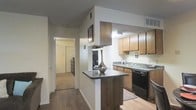 This single-floor residence offers a practical and comfortable layout designed for everyday living. The kitchen is a standout feature, showcasing warm wood cabinetry that provides ample storage space, a dark countertop breakfast bar that serves as both a workspace and casual dining area, and a full suite of appliances including a black dishwasher and electric range. The sink is positioned along the far counter, offering a functional workflow throughout the cooking space. A textured ceiling adds subtle character to the kitchen area. The flooring throughout is a rich hardwood-style surface that flows seamlessly from the kitchen into the hallway, creating a cohesive and visually connected space. The hallway extends toward additional rooms, with doors visible in the background that hint at well-defined private spaces such as bedrooms. Neutral wall tones keep the space feeling light and open, while recessed and overhead lighting ensures consistent illumination throughout. A small decorative vase on the breakfast bar adds a touch of personal style. Overall, the residence balances functionality with a warm, understated aesthetic that makes it an inviting and livable space.
