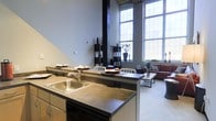 This two-floor loft space showcases a striking industrial aesthetic with soaring double-height ceilings and oversized multi-pane windows that flood the interior with abundant natural light. The open-concept main floor integrates the kitchen, dining, and living areas into one cohesive, flowing space. The kitchen features a central island with a dark countertop, a stainless steel double sink, and built-in appliances, making it both functional and visually prominent. Dark structural columns punctuate the space, adding to the industrial character while providing a bold contrast against the light-toned walls. The dining area sits adjacent to the kitchen island, creating a natural transition for entertaining. The living area is anchored by a comfortable seating arrangement positioned beneath the grand windows, taking full advantage of the natural light and open atmosphere. Neutral flooring ties the spaces together, contributing to the airy, loft-like ambiance. The two-floor layout offers additional versatility, providing a separation of living zones while maintaining the expansive, open feel that defines this space. The combination of raw industrial elements and modern finishes creates a dynamic and functional environment throughout.
