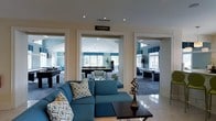 Located at 361 Darlington Avenue in Wilmington, NC, this single-floor space is designed for social engagement and leisure. The interior showcases a refined coastal-inspired aesthetic, with a cool blue and crisp white color scheme carried consistently throughout. The main lounge area features polished tile flooring that reflects the recessed lighting above, anchored by a plush teal sectional sofa adorned with patterned throw pillows and paired with a dark wood coffee table.

A clearly marked Game Room is accessible through wide, white-trimmed doorways that lend an open and connected feel to the layout. Inside, multiple billiard tables are arranged across a carpeted floor, surrounded by abundant natural light streaming in through several large windows framed with blue Roman shades. The vaulted ceiling in the game room adds a sense of vertical spaciousness to the area.

An additional adjoining space, visible through a second wide opening, offers further seating and entertainment options, maintaining the same polished blue-and-white design language. Throughout the space, the architectural detailing, generous window placement, and cohesive furnishings create an inviting and well-appointed environment suited for recreation and socializing.