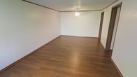 This single-floor space presents a clean, straightforward layout designed for practical living. The main living area is an open, generously proportioned room that immediately draws attention with its warm, medium-toned hardwood-style flooring, which runs throughout and adds a sense of warmth and continuity to the space. The walls are finished in a crisp white, providing a neutral backdrop that enhances the brightness of the room and offers flexibility for personalization. Crown molding trim lines the upper perimeter of the walls, adding a subtle architectural detail that elevates the overall finish of the space. A ceiling fan with a built-in light fixture is centrally mounted, ensuring both air circulation and ambient lighting throughout the room. Baseboards in a complementary wood tone frame the floor line, tying together the flooring and wall finishes cohesively. A doorway visible toward the back of the main room provides access to additional areas of the space, suggesting a connected and functional floor plan. Overall, the space conveys a clean, move-in-ready atmosphere with classic, understated finishes that are both durable and versatile.