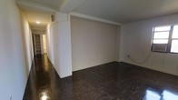 This single-floor space presents a straightforward, functional layout defined by its contrasting design elements. The most distinctive flooring feature is the dark parquet wood floor, which runs throughout the main area, adding a classic texture and depth to the space. The floors show a rich, dark tone with a traditional block pattern characteristic of parquet construction.

The walls are finished in a clean, neutral white, creating a bright contrast against the dark flooring. A prominent structural column is positioned centrally within the main open area, subtly dividing the space while maintaining an open feel. The ceiling is relatively modest in height and features a beam-like element along one side, adding subtle architectural detail.

A hallway extends toward the back of the space, leading to additional areas. Along the hallway, built-in shelving is visible, providing practical storage. Electrical outlets are positioned along the baseboards, and a wall-mounted unit is present near the upper portion of the wall, suggesting climate control capabilities.

Overall, the space offers a blank-canvas interior with neutral finishes, ready to be configured for a variety of uses.