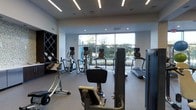 This fitness center offers a thoughtfully designed workout space that balances functionality with a bright, modern aesthetic. The room is finished with clean white walls and a neutral-toned floor, contributing to an open and energizing atmosphere. Overhead, a combination of linear LED lighting and recessed ceiling fixtures ensures consistent, well-distributed illumination throughout the space.

A standout feature is the large floor-to-ceiling window spanning the far wall, flooding the interior with natural light and providing an outward view of surrounding greenery and the exterior landscape — an inviting backdrop that enhances the workout environment.

The fitness center is equipped with a varied selection of cardio machines, including treadmills, ellipticals, and a stationary bike, all featuring individual digital display consoles. Additional workout equipment is positioned throughout the space to support a range of exercise routines. Along one wall, a dark shelving unit with a grid-style design provides organized storage, likely for towels or accessories, adding a practical touch to the overall layout. The space is efficiently arranged to allow comfortable movement between machines.