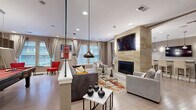 Located at 750 Summer St in Stamford, Connecticut, this well-appointed single-floor amenity space offers a thoughtfully designed layout that combines comfort and functionality. The open-plan living and lounge area serves as the centerpiece, showcasing a floor-to-ceiling natural stone fireplace surround with a wall-mounted flat-screen TV, recessed lighting, and a glass partition wall that adds a sleek architectural element. Plush seating, an area rug, and a glass coffee table create an inviting atmosphere, while large windows dressed with floor-length drapes allow natural light to fill the room. Adjacent to the lounge, the modern kitchen features dark wood cabinetry, granite-style countertops, a mosaic tile backsplash, stainless steel appliances, and contemporary pendant lighting — ideal for catering and casual use. The dining area offers a dedicated space with ample natural light from oversized windows overlooking the surrounding streetscape. A separate game room houses a billiards table alongside vibrant red accent seating and a dining set beneath bold artwork. The hallway connects all areas seamlessly, with light hardwood-style flooring running throughout and a neutral taupe color palette that lends a cohesive, modern aesthetic to the entire space.
