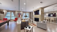 Located at 750 Summer St in Stamford, Connecticut, this well-appointed single-floor amenity space offers a thoughtfully designed layout that combines comfort and functionality. The open-plan living and lounge area serves as the centerpiece, showcasing a floor-to-ceiling natural stone fireplace surround with a wall-mounted flat-screen TV, recessed lighting, and a glass partition wall that adds a sleek architectural element. Plush seating, an area rug, and a glass coffee table create an inviting atmosphere, while large windows dressed with floor-length drapes allow natural light to fill the room. Adjacent to the lounge, the modern kitchen features dark wood cabinetry, granite-style countertops, a mosaic tile backsplash, stainless steel appliances, and contemporary pendant lighting — ideal for catering and casual use. The dining area offers a dedicated space with ample natural light from oversized windows overlooking the surrounding streetscape. A separate game room houses a billiards table alongside vibrant red accent seating and a dining set beneath bold artwork. The hallway connects all areas seamlessly, with light hardwood-style flooring running throughout and a neutral taupe color palette that lends a cohesive, modern aesthetic to the entire space.