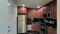 This single-floor space showcases a thoughtfully designed kitchen that serves as its centerpiece. The kitchen features rich, dark-toned raised-panel wood cabinetry that extends floor to ceiling, offering generous storage throughout. Complementing the cabinetry are sleek dark granite countertops paired with a matching black tile backsplash, creating a cohesive and elegant aesthetic. The kitchen is fully equipped with a suite of stainless steel appliances, including a full-size refrigerator, dishwasher, electric range, and an over-the-range microwave. Recessed lighting is strategically placed to illuminate the workspace effectively, while a stainless steel sink with a modern faucet adds a functional and polished touch. The dark tile flooring grounds the space and ties together the overall design palette. The layout is efficiently arranged in an L-shape configuration, maximizing both counter space and workflow. The overall ambiance of the kitchen is warm yet refined, combining classic cabinetry design with modern fixtures and finishes for a timeless, functional interior.