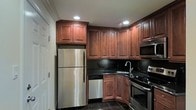 This single-floor space showcases a thoughtfully designed kitchen that serves as its centerpiece. The kitchen features rich, dark-toned raised-panel wood cabinetry that extends floor to ceiling, offering generous storage throughout. Complementing the cabinetry are sleek dark granite countertops paired with a matching black tile backsplash, creating a cohesive and elegant aesthetic. The kitchen is fully equipped with a suite of stainless steel appliances, including a full-size refrigerator, dishwasher, electric range, and an over-the-range microwave. Recessed lighting is strategically placed to illuminate the workspace effectively, while a stainless steel sink with a modern faucet adds a functional and polished touch. The dark tile flooring grounds the space and ties together the overall design palette. The layout is efficiently arranged in an L-shape configuration, maximizing both counter space and workflow. The overall ambiance of the kitchen is warm yet refined, combining classic cabinetry design with modern fixtures and finishes for a timeless, functional interior.