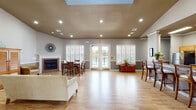 Located at 21300 Cotton Creek Dr in Gulf Shores, Alabama, this single-story community clubhouse offers a well-appointed, open-concept layout designed for comfortable gatherings and social events. The main gathering area is anchored by light hardwood flooring that flows throughout the space, neutral gray walls with contrasting white trim, and a raised tray ceiling with recessed lighting and a central skylight that floods the room with natural light. A classic fireplace serves as a focal point on one wall, while a set of French doors with flanking plantation-shuttered windows opens directly to an outdoor pool area, providing bright views and seamless indoor-outdoor access. The kitchen features warm honey-toned cabinetry, granite-style countertops, stainless steel appliances, and a peninsula bar with seating for multiple guests — ideal for catered events or casual use. A formal entry area showcases soaring ceilings and a pair of elegant white columns that frame the space, lending an architectural distinction to the welcoming foyer. Adjacent sitting areas offer versatile lounge space, with one featuring dramatically tall ceilings and generous wall space. Throughout, the design balances a coastal-casual aesthetic with functional, community-oriented amenities.
