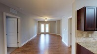 This single-floor space offers a clean, move-in-ready layout designed with both comfort and functionality in mind. The main living area is the centerpiece of the home, showcasing rich hardwood-style flooring that runs throughout and reflects the natural light that pours in through a set of classic French doors. These doors serve as a focal point, providing a bright, welcoming atmosphere while offering direct access to the outdoors. Neutral gray walls create a versatile backdrop that complements a wide range of furnishing styles, while crisp white baseboards and door trim add a polished, finished look. A ceiling fan with a light fixture is centrally positioned, ensuring year-round comfort and additional illumination. The open layout of the living space allows for flexible furniture arrangements and a generous sense of room. Adjacent doorways suggest additional rooms branching off the main area, hinting at a practical and well-organized floor plan. HVAC vents are visible both on the walls and floor, indicating a comprehensive climate control system. Overall, the space exudes a fresh, neutral aesthetic that is both inviting and easy to personalize.