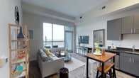 This contemporary single-floor residence offers a thoughtfully designed open-concept layout that seamlessly connects the kitchen, dining, and living areas. The living room is anchored by large floor-to-ceiling windows and a sliding glass door leading to a private balcony, flooding the space with natural light and offering sweeping urban skyline views. Durable wood-look flooring runs throughout the main living areas, complementing the modern aesthetic.

The kitchen is well-equipped with sleek gray flat-front cabinetry, dark granite countertops, a white tile backsplash, and a full suite of stainless steel appliances including a range, microwave, dishwasher, and refrigerator. A compact dining area adjacent to the kitchen provides a casual bar-height table with seating, ideal for everyday meals.

The bedroom is a calm, neutral retreat featuring plush carpeting, high ceilings, and a generous walk-in closet. A built-in dresser area with a wall mirror adds functional storage. The full bathroom is finished with clean white tile, a glass-enclosed walk-in shower, and light neutral flooring, maintaining a crisp and minimal aesthetic throughout. The overall design of the space is modern, functional, and urban.