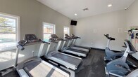 Located at 212 Catalpa Dr in Bloomington, IL, this single-floor fitness center is a dedicated exercise facility designed to support a full range of workout routines. The space is divided into two connected zones, separated by a distinctive dark-painted arched architectural element that adds visual interest while defining the areas.

The cardio zone features multiple treadmills, elliptical machines, and a stationary bike, all arranged to take advantage of the natural light streaming in through several large windows. A wall-mounted television is positioned for easy viewing during workouts. The bright, neutral-toned walls and dark rubber flooring throughout create a clean, functional aesthetic.

The strength training area houses a multi-station cable machine and a weight bench, catering to a variety of resistance exercises. A large floor-to-ceiling mirror spans one wall, enhancing the sense of space and allowing users to monitor their form. Recessed and overhead lighting ensures the facility is well-lit at all times.

Multiple entry and exit doors provide convenient access, and the open layout allows for comfortable movement between equipment. The overall design is straightforward and practical, prioritizing functionality and ease of use in a tidy, well-maintained environment.