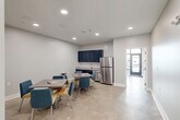 Located at 1717 Religious St in New Orleans, Louisiana, this single-floor communal dining space offers a clean, modern design well-suited for shared or multi-use environments. The layout is efficiently organized, combining a dining area with a functional kitchenette along one wall.

The kitchenette is equipped with navy blue upper cabinetry, a granite-topped counter with an undermount sink, a stainless steel dishwasher, and a full-size stainless steel refrigerator — providing all essential amenities in a compact footprint. The cabinetry's deep navy tones provide a striking contrast against the light gray walls.

The dining area accommodates multiple rectangular tables paired with a mix of blue upholstered and yellow accent chairs, creating a welcoming, contemporary seating arrangement. Polished concrete flooring runs throughout, adding an industrial yet refined character to the space.

Recessed ceiling lighting ensures consistent, even illumination across the entire area. The space features multiple doorways, including an entry with glazed panels that allow natural light to filter in from outside. Additional rooms are accessible through interior doors, suggesting flexible use potential. The overall ambiance is bright, neutral, and purposefully designed for communal gathering and everyday use.