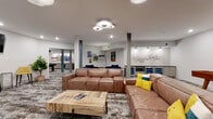 This single-floor amenity space is thoughtfully designed to serve as a dynamic social and recreation hub. The open-plan layout flows seamlessly between distinct activity zones, unified by a consistent palette of white walls, blue-gray accent walls, and patterned area rugs over hard flooring.

The main lounge area is anchored by a generously sized sectional and sofa in rich brown leather, arranged around a live-edge wood coffee table. A wall-mounted flat-screen TV and a row of yellow upholstered bar stools provide additional seating and entertainment options. Distinctive circular flush-mount light fixtures illuminate the space with a contemporary flair.

Adjacent to the lounge, the game room features two full-size pool tables — one with a slate-gray felt top and another with a classic black finish — each highlighted by large drum pendant lights overhead. A foosball table occupies a dedicated alcove with a blue accent wall, decorated with framed vintage-style artwork that adds character and personality to the space.

A quiet retreat nook furnished with two plush navy velvet armchairs, a side table, and a floor lamp offers a more intimate setting, adorned with a curated collection of travel and retro poster art. Toward the back, a wet bar area with cabinetry, under-counter beverage refrigerators, and display shelving provides convenient service for gatherings. Structural columns throughout are finished in blue-gray, contributing to the cohesive modern-industrial aesthetic of the overall space.