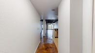 This single-floor space offers a compelling mix of industrial architecture and residential functionality, making it well-suited for live/work use or creative studio purposes. The most striking architectural feature is the soaring exposed steel ceiling with dark-painted corrugated metal decking and structural beams, which runs throughout the open main area, creating a dramatic loft-like atmosphere with generous vertical space.

Stained concrete floors extend across the majority of the space, showcasing rich, varied brown tones that complement the industrial aesthetic. The expansive main open area benefits from natural light through gridded windows, while a chandelier adds a contrasting residential warmth above the living zone. A half-wall peninsula separates the kitchen from the open area, maintaining visual connectivity throughout.

The kitchen is equipped with warm wood cabinetry, dark countertops, stainless steel appliances including a refrigerator, range, and microwave, and light ceramic tile flooring. Two bathrooms are present: one features a tiled tub/shower combo with a single-sink vanity, while the other offers a walk-in tiled shower with access to a walk-in closet. Additional rooms with stained concrete floors and tall ceilings provide flexible space. A hallway with textured white walls connects the private areas to the main open floor.