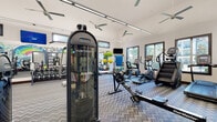 This single-level fitness center is designed to accommodate a wide range of workout styles and fitness goals. The main workout area is generously proportioned, featuring a patterned chevron flooring that extends throughout most of the space. It is outfitted with a comprehensive selection of equipment, including cable machines, a Nautilus Instinct multi-functional trainer, rowing machines, ellipticals, a stair climber, and a treadmill. A full rack of kettlebells and dumbbells lines one wall alongside floor-to-ceiling mirrors, enhancing the sense of openness. Large windows along the perimeter allow natural light to filter in and offer views of surrounding greenery, while ceiling fans and linear LED lighting maintain a comfortable, well-lit environment. Mounted televisions are positioned throughout for convenience during workouts.

A dedicated functional training annex features bright yellow flooring, TRX suspension straps, a weighted sled, a tire flip station, and a cable machine, set against mirrored walls and its own wall-mounted display. A bold, colorful mural depicting an Austin-themed scene with a vintage pickup truck, rainbow, and cactus serves as an eye-catching focal point near the entrance, adding a strong sense of local character and energy to the overall space. A built-in refreshment station with dark cabinetry is also available near the main entrance.