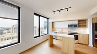 This well-appointed one-bedroom, one-bathroom unit offers a thoughtfully designed layout across a single floor, combining modern aesthetics with functional living spaces. The open-concept kitchen and living area serve as the heart of the home, bathed in natural light from large, black-framed floor-to-ceiling windows that frame expansive city and skyline views. The kitchen is outfitted with stainless steel appliances — including a range, microwave, and dishwasher — complemented by light wood cabinetry, white upper cabinets, and a herringbone-patterned tile backsplash. A peninsula with a white quartz countertop provides additional prep space and a casual dining option. Light wood flooring runs throughout, creating a warm and cohesive feel. The living area connects seamlessly to the kitchen, with an open passage leading to the private bedroom. The bedroom is a calm retreat featuring large windows with city views, a built-in closet with organized shelving, and the same warm hardwood flooring. The bathroom is clean and modern, featuring a glass-enclosed walk-in shower with white subway tile, a wood-toned vanity, and neutral stone-look tile flooring. A hallway provides access to additional storage and in-unit laundry, completing this efficient and stylish urban residence.