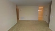 This single-story space presents a clean, open layout with a neutral and understated aesthetic throughout. The main living area is finished with wall-to-wall beige carpeting, which adds warmth underfoot and contributes to the space's calm, muted color palette. Smooth cream-colored walls are paired with white baseboards and trim, creating a simple yet cohesive interior environment.

A floor-level HVAC vent is centrally positioned along the main wall, indicating a built-in climate control system. A hallway visible on the left side of the living area provides access to additional rooms beyond, suggesting a functional and straightforward floor plan.

Adjacent to the living area, a dining space is partially visible to the right, distinguished by hardwood or laminate flooring and an elegant chandelier overhead. The chandelier features a classic multi-arm brass-toned design, lending a touch of traditional style to the otherwise understated interior.

Overall, the space offers a practical and move-in-ready environment with consistent finishes throughout, well-suited for those seeking a clean, neutral canvas to personalize.