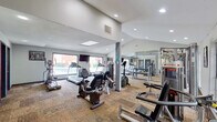 This fitness center is designed to support a comprehensive workout experience across two distinct areas. The main gym room features vaulted ceilings with recessed lighting and a skylight that brings in natural light, creating an open and energizing atmosphere. Large windows overlook an outdoor pool area, adding a bright, airy feel to the space. The room is outfitted with a wide selection of cardio equipment, including multiple treadmills, ellipticals, and stationary bikes, alongside a multi-station cable machine and additional strength-training apparatus. A wall-mounted flat-screen TV provides entertainment during workouts, and full-length mirrors along one wall visually expand the space while allowing users to monitor their form. The durable, patterned carpet tile flooring adds traction underfoot throughout this area. A separate studio room offers a clean, open layout with smooth hardwood-style flooring, ideal for yoga, stretching, dance, or group fitness classes. This room features a full mirrored wall, wall-mounted fans for ventilation, stability balls for functional training, and its own exterior window for natural light. Together, both areas provide a well-rounded and functional fitness environment suited to a variety of exercise routines.