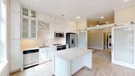 Located at 600 Caraway Crossing in Chapel Hill, NC, this thoughtfully designed single-floor residence offers a bright and cohesive living environment finished with light hardwood-style flooring, neutral tones, and crown molding throughout. The kitchen is a standout feature, showcasing floor-to-ceiling white cabinetry with glass-front uppers, a decorative herringbone tile backsplash, stainless steel appliances, and a mobile island with a clean white countertop that provides additional prep and serving space. A frosted glass barn door adds a modern, stylish touch that connects the kitchen to the living area, which is equipped with a ceiling fan and recessed lighting. Large windows and a glass-paneled balcony door flood the main living area with natural light while offering views of the surrounding Chapel Hill streetscape. The bedroom features a spacious layout with multiple closets, including a walk-in with wire shelving, and a built-in mudroom-style nook for added organization. The bathroom is finished with light tile flooring, a glass-enclosed shower, and a white vanity. An in-unit laundry area with a stacked washer and dryer is conveniently accessible from the bathroom. The space is completed by a carpeted room ideal for use as a secondary bedroom or flex space, featuring notably high ceilings.