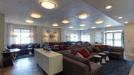 This single-floor lounge space offers an open, airy environment designed for comfort and social interaction. The layout is thoughtfully arranged with multiple seating clusters, each featuring large, cushioned gray sofas accented with bold red and patterned throw pillows, creating a warm and inviting ambiance throughout.

The ceiling is a standout architectural feature, adorned with a grid of round, drum-style light fixtures that cast a warm, even glow across the entire space. These fixtures are complemented by wall-mounted sconces and freestanding floor lamps, ensuring the room is well-lit from multiple levels.

Along the perimeter, a series of windows dressed with sheer white curtains allow natural light to filter softly into the interior, enhancing the bright and open feel of the space. Decorative starburst wall accents add a stylish, contemporary touch to the white walls between the windows.

The flooring is a sleek, dark surface that contrasts elegantly with the lighter tones of the furniture and walls. White side tables and a central credenza provide functional surfaces while maintaining the clean, modern aesthetic. The overall design balances a sophisticated, neutral palette with energetic pops of color, making this a versatile and visually engaging space suited for relaxation or social gatherings.