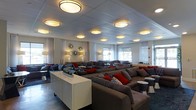 This single-floor lounge space offers an open, airy environment designed for comfort and social interaction. The layout is thoughtfully arranged with multiple seating clusters, each featuring large, cushioned gray sofas accented with bold red and patterned throw pillows, creating a warm and inviting ambiance throughout.

The ceiling is a standout architectural feature, adorned with a grid of round, drum-style light fixtures that cast a warm, even glow across the entire space. These fixtures are complemented by wall-mounted sconces and freestanding floor lamps, ensuring the room is well-lit from multiple levels.

Along the perimeter, a series of windows dressed with sheer white curtains allow natural light to filter softly into the interior, enhancing the bright and open feel of the space. Decorative starburst wall accents add a stylish, contemporary touch to the white walls between the windows.

The flooring is a sleek, dark surface that contrasts elegantly with the lighter tones of the furniture and walls. White side tables and a central credenza provide functional surfaces while maintaining the clean, modern aesthetic. The overall design balances a sophisticated, neutral palette with energetic pops of color, making this a versatile and visually engaging space suited for relaxation or social gatherings.