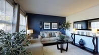 This single-floor space makes a striking first impression with its thoughtfully designed living area. The focal point of the room is a deep navy blue accent wall, which provides a bold yet refined backdrop for the neutral-toned seating arrangement. A plush, light-colored sofa adorned with decorative patterned pillows anchors the space, paired with a sleek round glass-top coffee table supported by a dark wood base — a combination that balances comfort with sophistication.

Wall-mounted black candle sconce fixtures flank a pair of framed landscape art pieces on the accent wall, adding a classic decorative touch. A dark console table along the adjacent wall holds a warm-glowing lamp, contributing to the cozy lighting atmosphere alongside a matching table lamp on the opposite side of the room.

Natural light filters in through a window dressed with layered curtains, brightening the white ceiling and lighter walls that contrast elegantly with the navy feature wall. Lush indoor plants are positioned throughout the room, introducing organic texture and a refreshing natural element. A soft, thick area rug grounds the seating area, enhancing the overall warmth and comfort of the space. The room's design reflects a cohesive, polished aesthetic with a careful balance of dark and light tones.
