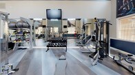 Located at 10001 E Dry Creek Rd in Englewood, Colorado, this fitness center is a thoughtfully designed, single-floor facility built to support a comprehensive workout experience. The space is defined by its impressively high ceilings, which lend an open and airy atmosphere, complemented by large windows that allow generous natural light to fill the room. A bold two-tone color scheme of navy blue and neutral cream runs throughout, accented by teal tile detailing and large motivational fitness imagery mounted on the walls.

The flooring consists of wide-plank hardwood-style material that adds a clean, modern aesthetic across the entire space. A full wall of mirrors along one side visually expands the room while providing functional feedback for strength training. Overhead, a large ceiling fan ensures consistent air circulation during workouts.

The cardio zone is well-stocked with Cybex treadmills, elliptical trainers, stationary bikes, a rowing machine, and a spin bike, each positioned to take advantage of the natural light from the surrounding windows. Multiple flat-screen televisions are mounted throughout for entertainment. The strength area features a complete dumbbell rack and a multi-function cable machine, offering a full range of resistance training options. The layout provides ample open floor space, allowing for ease of movement between equipment zones.