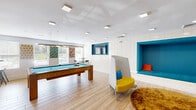 Located at 325 Commandants Way in Chelsea, Massachusetts, this café and lounge space offers a dynamic and well-appointed recreational environment designed for both socializing and relaxation. The open-plan layout is anchored by light wood-look flooring that runs throughout, lending warmth and continuity to the space.

A full-size billiards table serves as the centerpiece, accompanied by a dedicated cue rack mounted on a bold teal accent wall, along with a high-top table and stools for spectators. Natural light pours in through large windows on one end, brightening the entire area and offering views of the surrounding exterior.

On the opposite side, a built-in teal upholstered alcove provides a cozy, lounge-style seating nook, accented with colorful throw pillows. Modern high-back pod chairs in yellow and white are arranged nearby, alongside a plush round area rug, creating an inviting space for casual conversation or quiet moments. A wall-mounted flat-screen television is also integrated into the white shiplap feature wall.

Adjacent areas include a transitional corridor with glass partitions leading to additional amenity spaces, as well as a teal accent display shelf area. Recessed and flush-mount ceiling lighting throughout ensures the space remains bright and welcoming at all hours. The overall ambiance is modern, playful, and thoughtfully designed for community engagement.