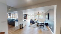 Located at 4040 26th Ave SW in Seattle, WA, this thoughtfully designed single-floor space offers a seamless open-plan layout that connects multiple living and entertaining zones with ease. Light hardwood flooring runs throughout, lending warmth and continuity to the entire space.

The main living area is anchored by a dramatic floor-to-ceiling brick fireplace wall, complete with a mounted flat-screen television, creating a striking focal point. Large floor-to-ceiling windows and a glass door leading to a private balcony flood the room with natural light and offer views of the surrounding Seattle neighborhood and lush tree canopy. A plush sectional sofa, paired with round wood accent tables atop a vibrant orange area rug, completes the inviting atmosphere.

A secondary lounge area features a cozy sectional arrangement beneath a decorative chandelier, with an accent wall in deep slate blue and its own wall-mounted television — ideal for a more intimate gathering space.

The kitchen is a standout feature, showcasing vivid cobalt blue countertops, a bold green subway tile backsplash, stainless steel appliances including a double wall oven, open shelving, and a generous island with bar seating illuminated by industrial-style pendant lights.

Recessed lighting, an elegant chandelier, and carefully chosen furnishings throughout contribute to a modern, polished ambiance suited for both relaxation and entertaining.