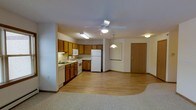 This single-floor space is designed with an open-concept layout that connects the kitchen and living area in a seamless, functional arrangement. The kitchen is well-appointed with warm honey-toned wood cabinetry offering generous storage, a white appliance suite including a refrigerator, range, dishwasher, and built-in microwave, and a sink positioned beneath under-cabinet lighting. Light hardwood flooring runs throughout the kitchen and main living area, transitioning to plush carpet in the designated living zone, creating a subtle yet defined separation of spaces. A ceiling fan with an integrated light fixture is centrally positioned overhead, providing both airflow and ambient lighting. A pendant light hangs near the entryway area, adding a welcoming accent. The entry door, finished in a natural wood tone, opens directly into the main living space, offering convenient access. Neutral-toned walls and consistent, bright overhead lighting contribute to a clean and airy atmosphere throughout. The layout is practical and efficient, making excellent use of the available floor plan to create a cohesive, comfortable living environment.