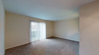 This single-floor residence offers a practical and cohesive layout across multiple rooms, unified by neutral beige tones and consistent design elements throughout. The living area is a spacious, open room finished with plush wall-to-wall carpeting and features a sliding glass door dressed with vertical blinds, allowing generous natural light to filter in while providing direct access to an outdoor space.

The kitchen is a functional galley-style space featuring white cabinetry that extends along one wall, offering ample storage both above and below the counter. Light-toned countertops provide a clean workspace, while a stainless steel sink sits beneath under-cabinet lighting. White appliances, including a range and dishwasher, complement the cabinetry, and durable wood-look flooring grounds the room. A pass-through window connects the kitchen visually to the adjacent living area.

The bedrooms are carpeted and finished with the same warm neutral palette found throughout the unit. Each room is bright, with windows fitted with blinds offering views of the surrounding neighborhood. One bedroom includes dual doors, suggesting a closet alongside a hallway entrance, adding practical storage. Clean white baseboards and trim are consistent throughout, contributing to a tidy, move-in ready ambiance.