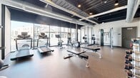Located at 10 King Street in Troy, NY, this fitness center offers a thoughtfully designed exercise space that balances industrial aesthetics with functional fitness amenities. The room features an exposed dark ceiling with visible ductwork and structural beams, lending the space a modern, urban character. Durable rubber flooring in a warm brown tone covers the entire area, providing traction and cushioning throughout.

A defining architectural feature is the floor-to-ceiling glass storefront wall facing the street, which floods the room with abundant natural light and offers open views of the surrounding King Street neighborhood. This transparent facade creates an energizing, connected atmosphere for workouts.

The exercise room is comprehensively equipped for a full-body training experience. Cardio equipment includes multiple treadmills, an elliptical trainer, and a stationary bike, all positioned to take advantage of the street views. Strength training is well represented with a multi-function cable machine, a power rack with Olympic weight plates, adjustable weight benches, and two sets of dumbbell racks offering a wide range of weights. Kettlebells, stability balls, and floor mats round out the functional fitness offerings. A large wall-mounted mirror in the free weights area enhances the sense of space and supports proper form during training sessions.