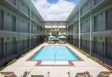 Located in Metairie, Louisiana, this single-floor unit is part of a multi-story residential complex centered around a large, fenced outdoor pool courtyard. The courtyard features a rectangular swimming pool surrounded by a brick-trimmed pool deck, lounge chairs, and shaded umbrella seating, all framed by symmetrical building facades with open-air corridors and balconies.

Inside, the living area offers a warm, comfortable atmosphere with soft carpet flooring and smooth walls. A plush loveseat anchors the space, accompanied by decorative wall art and accent shelving. The layout transitions naturally into an adjacent area with hardwood-style flooring, where a bar-height table and stools provide a casual seating option.

The bedroom is thoughtfully arranged, featuring carpeted flooring, neutral-toned walls, and understated décor including framed artwork and a decorative ladder accent. A warm lamp and wooden dresser add to the room's welcoming character.

A transitional hallway area features rich tile flooring in a herringbone pattern, a sculptural table lamp, and a decorative round mirror flanked by indoor greenery, creating a polished entryway feel. The overall interior presents a cohesive, quietly elegant aesthetic with practical, well-considered design throughout.