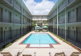 Located in Metairie, Louisiana, this single-floor unit is part of a multi-story residential complex centered around a large, fenced outdoor pool courtyard. The courtyard features a rectangular swimming pool surrounded by a brick-trimmed pool deck, lounge chairs, and shaded umbrella seating, all framed by symmetrical building facades with open-air corridors and balconies.

Inside, the living area offers a warm, comfortable atmosphere with soft carpet flooring and smooth walls. A plush loveseat anchors the space, accompanied by decorative wall art and accent shelving. The layout transitions naturally into an adjacent area with hardwood-style flooring, where a bar-height table and stools provide a casual seating option.

The bedroom is thoughtfully arranged, featuring carpeted flooring, neutral-toned walls, and understated décor including framed artwork and a decorative ladder accent. A warm lamp and wooden dresser add to the room's welcoming character.

A transitional hallway area features rich tile flooring in a herringbone pattern, a sculptural table lamp, and a decorative round mirror flanked by indoor greenery, creating a polished entryway feel. The overall interior presents a cohesive, quietly elegant aesthetic with practical, well-considered design throughout.
