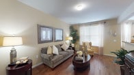 This single-floor residence offers a thoughtfully designed layout that balances comfort and functionality across several well-defined living spaces. The living room features warm hardwood-style flooring, soft neutral wall tones, and large windows dressed with sheer curtains that allow natural light to fill the room, creating an inviting and airy atmosphere.

Adjacent to the living area, a curved bay window distinguishes the dining space, where multiple windows bathe the room in natural light. The hardwood-style flooring continues seamlessly from the living area, reinforcing a cohesive aesthetic throughout the common areas.

The kitchen is well-equipped with rich warm-toned cabinetry extending to the upper walls, generous granite-style countertops, and black appliances including a refrigerator, electric range, built-in microwave, and dishwasher. Ample upper and lower cabinet storage provides practical functionality.

The bedroom offers a comfortable retreat with plush carpeting, a ceiling fan, and warm neutral walls. It connects to a spacious walk-in closet with full-width wire shelving and hanging rods. The bathroom features a wide vanity with extended countertop space, a large mirror, and hardwood-style flooring.

Completing the layout, a dedicated in-unit laundry room accommodates a full-sized washer and dryer alongside overhead shelving and hanging storage.