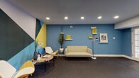 This single-floor fitness facility is thoughtfully designed to accommodate a full range of exercise needs within a cohesive, modern aesthetic. The space is united by a consistent color palette of steel blue accent walls, crisp white trim, and durable speckled rubber flooring that flows through each zone.

The welcoming lounge area makes an immediate visual impression with a bold geometric mural in teal, yellow, black, and white. Furnished with a cream loveseat, mid-century modern accent chairs, and a round wood coffee table, this area provides a comfortable space to relax before or after a workout. Decorative shelving, framed art, and a floor lamp add personality to the blue feature wall.

The fitness area is divided into several well-organized zones. A cardio section houses multiple treadmills, elliptical trainers, and a stationary bike, complemented by a wall-mounted television and natural light from slatted windows. A dedicated strength training zone features cable machines, a weight bench, a squat rack, and a full dumbbell rack set against floor-to-ceiling mirrors that visually expand the space. Wall-mounted yoga mats and stability balls signal a dedicated flexibility and stretching area. Recessed lighting throughout ensures consistent illumination across all workout zones.
