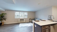 This single-floor unit offers a thoughtfully designed layout that maximizes functionality within a compact footprint. The open-plan living and kitchen area forms the heart of the space, where durable wood-look flooring flows seamlessly throughout. The living area is bathed in natural light from a large multi-pane window and features a wall-mounted HVAC unit, neutral warm-toned walls, and ample room for comfortable furnishings. A dedicated media corner with a TV stand and decorative indoor plant adds a homey touch.

The kitchen is efficiently arranged in a galley style, featuring white shaker cabinetry with modern bar-pull hardware, light countertops, and a full suite of stainless steel appliances including an electric range, over-the-range microwave, and refrigerator. A dark-finished kitchen island with a white countertop serves as both a prep area and casual dining surface, bridging the kitchen and living spaces.

The bathroom is clean and contemporary, showcasing a white shaker-style vanity with a large mirror, modern light fixture, and wood-look flooring that ties into the rest of the unit. The overall aesthetic is modern and neutral, creating a bright, cohesive living environment.