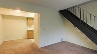 This two-story space offers a straightforward and functional layout across both levels. The main living area is finished with wall-to-wall carpet in a neutral beige tone, providing a comfortable and cohesive foundation throughout. White walls keep the interior feeling clean and open, while a floor vent indicates a forced-air climate system built into the home.

A staircase with dark metal railings rises along one wall, connecting the two floors and serving as a subtle architectural detail within the living space. The angled underside of the staircase adds visual structure to the room.

An adjacent kitchen is visible through a doorway opening, featuring wood-toned cabinetry in a warm honey finish, light countertops, and tile flooring that contrasts with the carpeted living area. Overhead lighting illuminates the kitchen, which includes base and upper cabinets offering practical storage. A door within the kitchen leads to an additional area of the unit.

Overall, the space presents a simple, utilitarian design with consistent neutral finishes, making it a versatile and adaptable environment suited to a range of uses and personal styles.