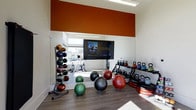 This fitness center is thoughtfully designed to accommodate a broad range of exercise needs in a clean, modern environment. The space features hardwood-style flooring and crisp white walls accented by a bold terracotta-orange upper wall panel, lending the room a vibrant and energizing atmosphere. A large wall-to-wall mirror expands the sense of space and allows natural light from the adjacent window to flow throughout the room. Mounted centrally on the mirror is a flat-screen TV, providing entertainment or instructional content during workouts. The facility is well-stocked with a comprehensive selection of equipment, including a tiered medicine ball rack with a full range of weighted balls, a multi-tier kettlebell rack with various sizes, stability and exercise balls in assorted sizes and weights, and stationary cycling equipment visible in the reflected mirror. Additional workout accessories, such as balance boards, are also present. The layout is open and functional, offering ample floor space for stretching, bodyweight exercises, or equipment-based training. Overall, the space provides a professional and inviting environment suited for both casual and dedicated fitness routines.