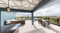 This single-floor space combines contemporary design with functional amenities across several distinct areas. The standout feature is a generously sized covered outdoor terrace, ideal for gatherings and relaxation. The terrace is finished with large-format porcelain tile flooring and is partially sheltered by a striking coffered ceiling with a geometric, paneled pattern that adds architectural interest overhead. Glass-panel railings line the perimeter, maintaining unobstructed panoramic views of the surrounding landscape, including lush tree canopies and open skies. Wicker lounge seating and dining sets furnish the space, creating distinct zones for socializing and dining al fresco.

Inside, the interiors are finished in a clean, neutral palette with light gray walls and coordinated gray trim throughout. A notable decorative focal point is an eye-catching wall-mounted sculptural installation—a cluster of organic, disc-shaped metallic elements that casts dramatic shadows, lending an artistic, gallery-like quality to the space. The corridors are carpeted in a light neutral tone and lead to ADA-accessible restroom facilities, clearly marked and finished with dark-framed glass doors, reflecting a commitment to accessibility and polished commercial design standards throughout.