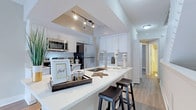This two-story residence is thoughtfully designed with a clean, contemporary aesthetic carried throughout both levels. The kitchen serves as a central highlight, showcasing white shaker cabinetry, a generous peninsula with an undermount sink and bar seating, stainless steel appliances including a refrigerator, microwave, range, and dishwasher, and bright white countertops. Track lighting above the peninsula adds a modern touch, while wood-look flooring extends through the main living areas, complementing the neutral color palette.

A carpeted staircase with white balusters connects the two floors, providing a classic contrast to the hard flooring below. The in-unit laundry area is conveniently tucked behind a paneled door and includes a full-size washer and dryer with overhead shelving.

The bathrooms are well-appointed, featuring white vanities with undermount sinks, quartz-style countertops, and large mirrors with globe lighting fixtures. One bathroom offers a corner vanity layout with dual sink areas, while another includes a deep soaking tub set within white tile surround with a small window above for natural light. Bedrooms are finished with wood-look flooring, neutral walls, and windows providing natural light.