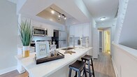 This two-story residence is thoughtfully designed with a clean, contemporary aesthetic carried throughout both levels. The kitchen serves as a central highlight, showcasing white shaker cabinetry, a generous peninsula with an undermount sink and bar seating, stainless steel appliances including a refrigerator, microwave, range, and dishwasher, and bright white countertops. Track lighting above the peninsula adds a modern touch, while wood-look flooring extends through the main living areas, complementing the neutral color palette.

A carpeted staircase with white balusters connects the two floors, providing a classic contrast to the hard flooring below. The in-unit laundry area is conveniently tucked behind a paneled door and includes a full-size washer and dryer with overhead shelving.

The bathrooms are well-appointed, featuring white vanities with undermount sinks, quartz-style countertops, and large mirrors with globe lighting fixtures. One bathroom offers a corner vanity layout with dual sink areas, while another includes a deep soaking tub set within white tile surround with a small window above for natural light. Bedrooms are finished with wood-look flooring, neutral walls, and windows providing natural light.