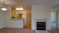 This single-floor space presents an open-concept layout that connects the living area and kitchen, creating a cohesive and functional flow throughout. The living area is anchored by a notable fireplace featuring a white tile surround and hearth, serving as a natural focal point within the room. Carpeting covers the living area floor, adding warmth and comfort underfoot.

The kitchen is positioned adjacent to the living space, separated by a raised breakfast bar or counter that provides both a visual divide and additional surface space. Kitchen cabinetry in a light wood tone lines the upper walls, accompanied by essential appliances including a microwave and refrigerator. Overhead fluorescent lighting illuminates the kitchen area effectively. A six-panel door is visible toward the back of the space, likely leading to an additional room or exterior access point.

The overall color palette is neutral, with white and beige tones throughout the walls, cabinetry, and fixtures, lending the space a clean and understated appearance. The layout is practical and compact, making efficient use of the available footprint across its single level.