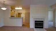 This single-floor space presents an open-concept layout that connects the living area and kitchen, creating a cohesive and functional flow throughout. The living area is anchored by a notable fireplace featuring a white tile surround and hearth, serving as a natural focal point within the room. Carpeting covers the living area floor, adding warmth and comfort underfoot.

The kitchen is positioned adjacent to the living space, separated by a raised breakfast bar or counter that provides both a visual divide and additional surface space. Kitchen cabinetry in a light wood tone lines the upper walls, accompanied by essential appliances including a microwave and refrigerator. Overhead fluorescent lighting illuminates the kitchen area effectively. A six-panel door is visible toward the back of the space, likely leading to an additional room or exterior access point.

The overall color palette is neutral, with white and beige tones throughout the walls, cabinetry, and fixtures, lending the space a clean and understated appearance. The layout is practical and compact, making efficient use of the available footprint across its single level.