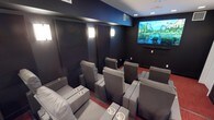 This purpose-built home theater is thoughtfully designed to deliver a true cinematic experience. The room is finished in deep charcoal tones throughout, with dark walls and a light ceiling that draw focus to the large wall-mounted display screen positioned at the front of the space. Rows of plush gray leather reclining seats are arranged in a tiered, stadium-style layout, ensuring unobstructed sightlines from every position. Each seat features built-in armrests and integrated cup holders, combining comfort with convenience for extended viewing sessions. The flooring is finished in a rich red carpet that adds warmth and contrast to the otherwise dark palette, while also contributing to the room's acoustic performance. Along the side and rear walls, large acoustic panels are mounted between elegant wall sconces, which provide soft, ambient illumination without disrupting the viewing atmosphere. Recessed ceiling lights offer additional controlled lighting, and a dedicated ventilation system ensures year-round comfort. A soundbar is mounted beneath the screen, complementing the room's audio setup. The space is accessed through a solid door and is designed to minimize light and sound intrusion, creating a fully immersive, distraction-free environment ideal for film enthusiasts and entertainment alike.