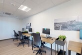 Located at 6775 N 67th Ave in Glendale, Arizona, this single-room office space offers a well-organized and efficiently designed work environment. The layout accommodates multiple workstations arranged in a row, each equipped with computer monitors, ergonomic task chairs, and wood-toned desks that contribute to a warm yet professional atmosphere.

The interior features light wood-look flooring throughout, crisp white walls, and a suspended ceiling with recessed panel lighting that ensures consistent, even illumination across the workspace. A glass-panel door and adjacent glazed sidelight provide a visual connection to the adjacent corridor or suite, allowing natural light to filter into the room and enhancing the sense of openness.

Decorative touches include black-and-white photographic wall art and small potted plants placed between workstations, adding character and a touch of greenery to the otherwise clean and minimal design. The ceiling incorporates a visible HVAC unit, ensuring climate control suited to Arizona's warm climate.

The overall ambiance is modern, clean, and functional — designed to support focused, productive work. The space is ideal for a small team or shared office setting, with its thoughtful layout making efficient use of the available footprint while maintaining a comfortable and inviting work environment.