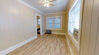 This single-floor space showcases a clean, functional layout with notable classic architectural details. The main living area features light-toned hardwood-style flooring that runs throughout, lending warmth and continuity to the space. Crown molding frames the ceiling, adding a refined touch to the otherwise simple design. A textured ceiling with a central ceiling fan provides both air circulation and ambient lighting.

Natural light enters through multiple windows fitted with blinds, including a double window at the far wall that overlooks an exterior view. Climate control is handled by a cast-iron radiator positioned beneath the double window, complemented by a wall-mounted air conditioning unit near the side window — a combination suited for year-round comfort.

The neutral, warm beige walls are accented by crisp white baseboards and door trim, creating a balanced and inviting atmosphere. A doorway visible from the main area leads into an adjacent room, suggesting a bedroom nearby, offering a sense of connected, practical flow between spaces. Overall, the unit presents a tidy, well-maintained environment with functional amenities and timeless detailing.