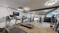 Located at 8500 Tree Top Ct S in Miamisburg, Ohio, this fitness center offers a comprehensive workout facility within a single, efficiently organized space. The room features neutral-toned walls, dark wood accent beams along the ceiling, and durable patterned carpet flooring with a multi-toned striped design that adds visual texture underfoot.

The cardio section is well-appointed, housing multiple treadmills and a stationary bike, complemented by a wall-mounted flat-screen television for entertainment during workouts. Adjacent to the cardio area, a dedicated strength training zone includes multiple selectorized cable and resistance machines suited for a full-body routine. A stair climber and rowing machine round out the aerobic options.

The free weight area features a tiered dumbbell rack stocked with a range of weights, alongside medicine balls and stability equipment for functional fitness training. Large mirrors span a significant portion of the walls, enhancing the sense of openness and allowing users to monitor their form throughout their workout.

Overhead, flush-mounted light fixtures and track lighting provide consistent, bright illumination throughout the space. A dedicated entry door with a glass panel offers easy access. Overall, the fitness center is thoughtfully arranged to accommodate a variety of training styles in a clean, functional environment.