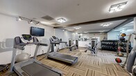 Located at 8500 Tree Top Ct S in Miamisburg, Ohio, this fitness center offers a comprehensive workout facility within a single, efficiently organized space. The room features neutral-toned walls, dark wood accent beams along the ceiling, and durable patterned carpet flooring with a multi-toned striped design that adds visual texture underfoot.

The cardio section is well-appointed, housing multiple treadmills and a stationary bike, complemented by a wall-mounted flat-screen television for entertainment during workouts. Adjacent to the cardio area, a dedicated strength training zone includes multiple selectorized cable and resistance machines suited for a full-body routine. A stair climber and rowing machine round out the aerobic options.

The free weight area features a tiered dumbbell rack stocked with a range of weights, alongside medicine balls and stability equipment for functional fitness training. Large mirrors span a significant portion of the walls, enhancing the sense of openness and allowing users to monitor their form throughout their workout.

Overhead, flush-mounted light fixtures and track lighting provide consistent, bright illumination throughout the space. A dedicated entry door with a glass panel offers easy access. Overall, the fitness center is thoughtfully arranged to accommodate a variety of training styles in a clean, functional environment.